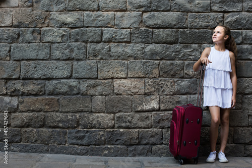 cheerful little girl traveler with suitcase leaning against stone wall of European city