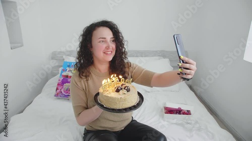 Young girl on a bed in a hotel, communicates and celebrates a birthday online and blows out candles on a cake