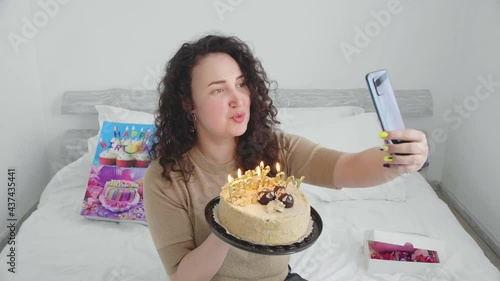 Young girl on a bed in a hotel, communicates and celebrates a birthday online and blows out candles on a cake