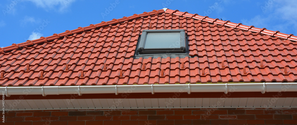 Naklejka premium Roofing construction. A red tiled roof with a skylight window installed, a plastic rain gutter, a fascia board and a soffit.