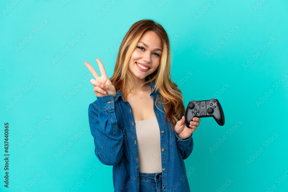 Teenager blonde girl playing with a video game controller over isolated wall smiling and showing victory sign Teenager blonde girl playing with a video game controller over isolated wall smiling and showing victory sign