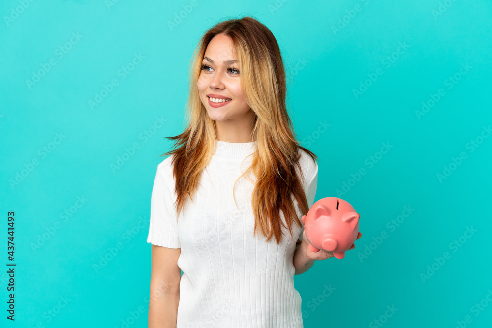 Teenager blonde girl holding a piggybank over isolated blue background looking side Teenager blonde girl holding a piggybank over isolated blue background looking side