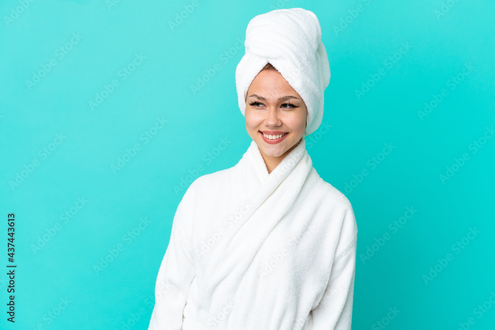 Teenager blonde girl in a bathrobe over isolated blue background looking to the side and smiling Teenager blonde girl in a bathrobe over isolated blue background looking to the side and smiling