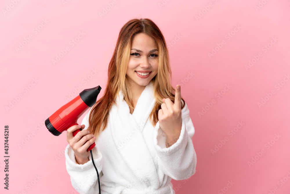 Teenager blonde girl holding a hairdryer over isolated pink background doing coming gesture