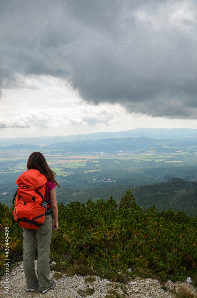 Back view of tourist girl with red backpack standing on top and enjoying breathtaking view to the valley. Tourism and travel concept.