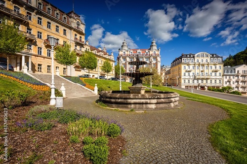 Marianske Lazne, Czech Republic - May 30 2021: The baroque water fountain standing on Goethe square. Green lawn, flowers and hotel buildings around. Sunny day with blue sky and clouds in spa city.