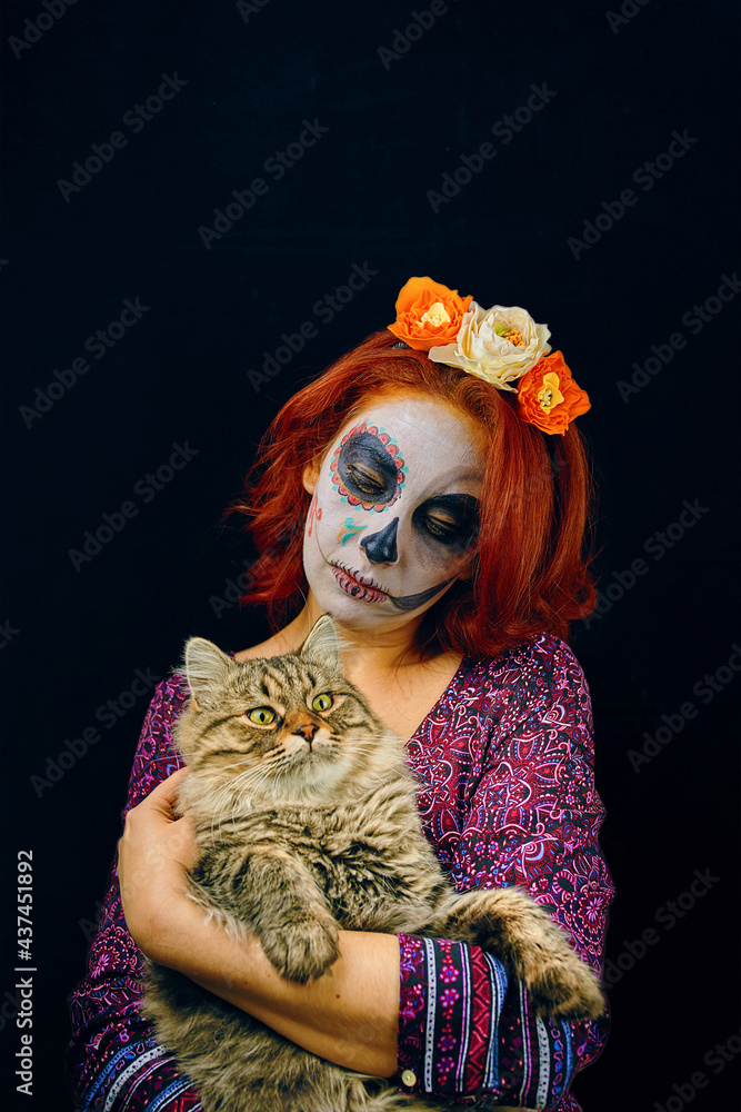 A young woman in day of the dead mask skull face art. Woman with skull makeup and red hair with cat in hands on dark background A young woman in day of the dead mask skull face art. Woman with skull makeup and red hair with cat in hands on dark background