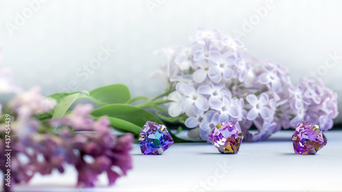 three lilac triangular crystals on a white table lilac flowers background, with a blurred branch of lilac in the foreground 