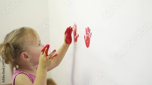 Little beautiful girl paints her handprints with red paint on a white wall.Slow motion.