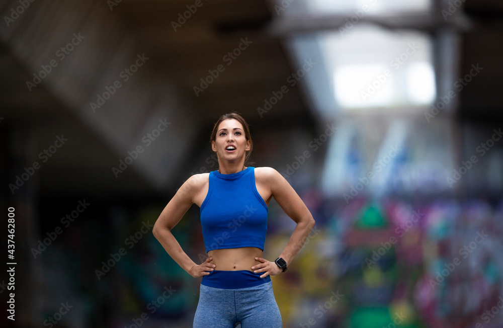 Female athlete standing with hands on hips breathing after workout in urban setting.