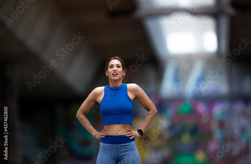 Female athlete standing with hands on hips breathing after workout in urban setting.