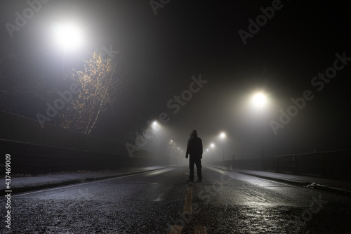 Low angle  looking up at a hooded figure standing in the middle of the road on an atmospheric foggy winters night. UK.