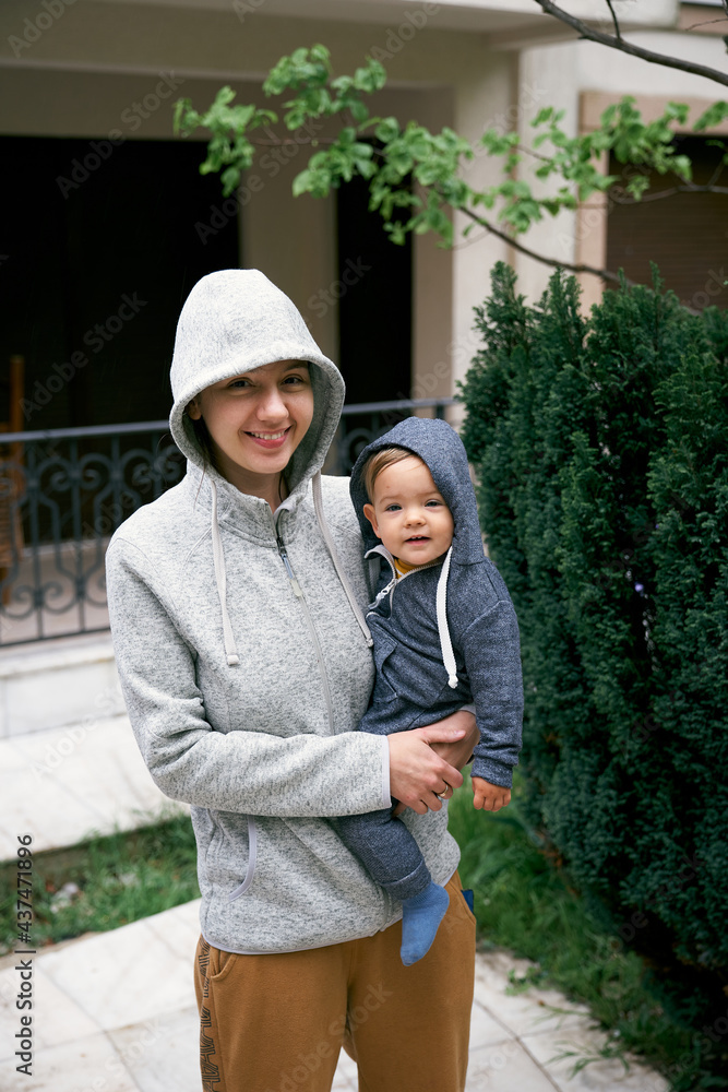 Smiling mom in a sweater with a hood holds a baby in overalls in her arms while standing in the courtyard of the house