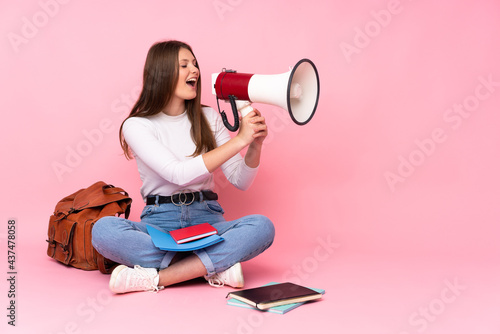 Teenager caucasian student girl sitting on the floor isolated on pink background shouting through a megaphone