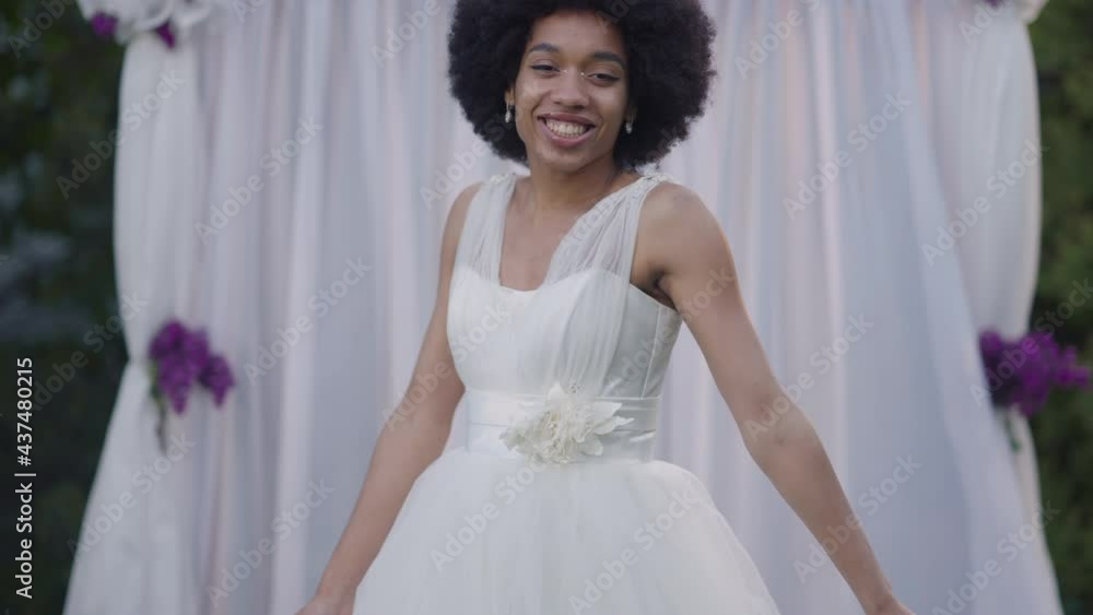 Front view of positive slim beautiful African American woman in white wedding dress jumping in slow motion with altar at background. Portrait of happy excited bride getting married outdoors Front view of positive slim beautiful African American woman in white wedding dress jumping in slow motion with altar at background. Portrait of happy excited bride getting married outdoors