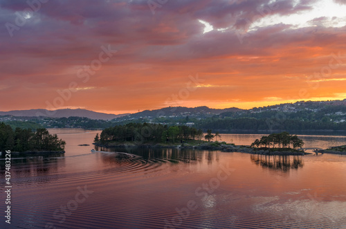 Colorful glow of a setting sun reflecting on the water as a boat glides between islands in a Norwegian landscape. 