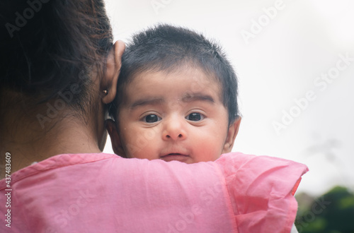 Cute baby girl peeking out from mothers shoulders with eyes wide open, looking at camera. Two months old sweet daughter with black hair hugging close to the warmth of her mom.