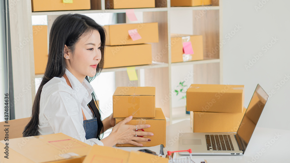 Asian businesswoman owning SME online using laptop and mobile phone. Receive customer orders with parcel boxes at her home office. online business ideas Asian businesswoman owning SME online using laptop and mobile phone. Receive customer orders with parcel boxes at her home office. online business ideas