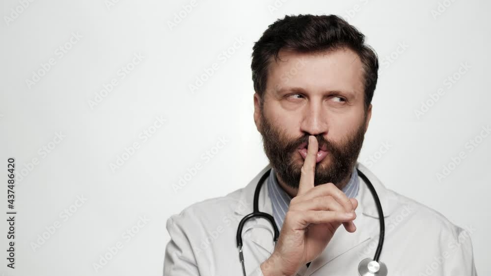 Shh, doctor secret finger. Suspicious smiling man doctor on white background looking at camera and brings his index finger to his mouth lips and she say shhh Shh, doctor secret finger. Suspicious smiling man doctor on white background looking at camera and brings his index finger to his mouth lips and she say shhh