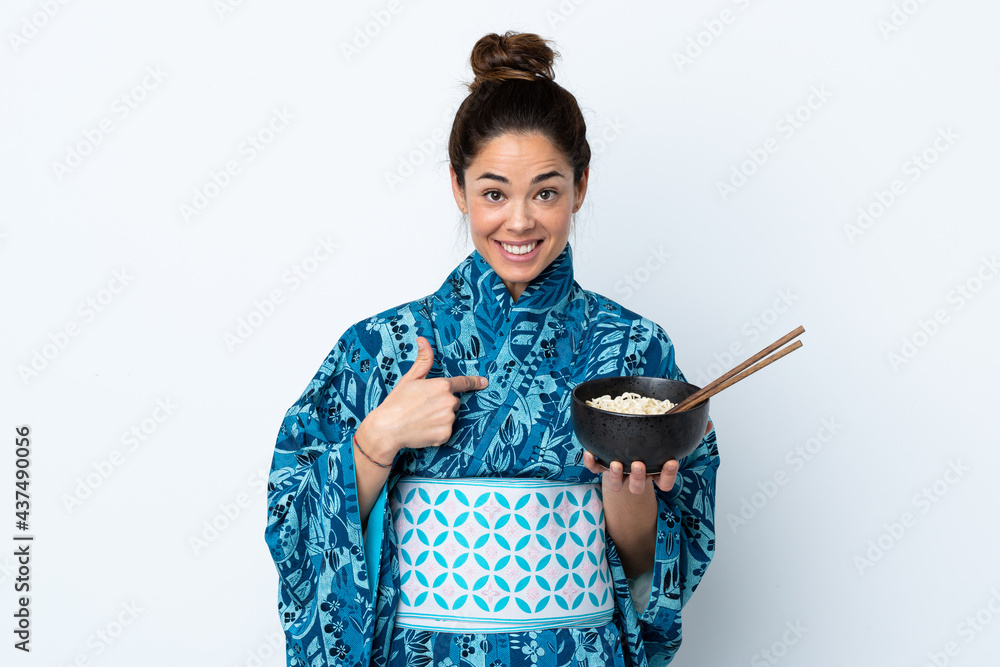Woman wearing kimono over isolated white background with surprise facial expression while holding a bowl of noodles with chopsticks Woman wearing kimono over isolated white background with surprise facial expression while holding a bowl of noodles with chopsticks