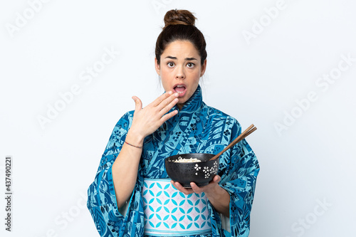 Woman wearing kimono over isolated white background holding a bowl of noodles with chopsticks and surprised