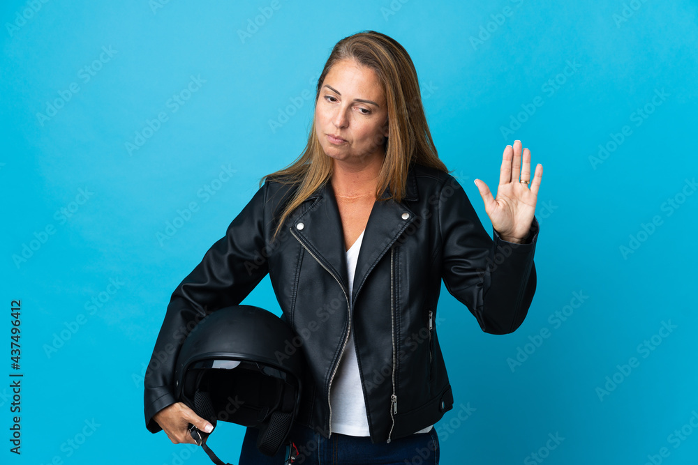 Middle age woman holding a motorcycle helmet isolated on blue background making stop gesture and disappointed Middle age woman holding a motorcycle helmet isolated on blue background making stop gesture and disappointed