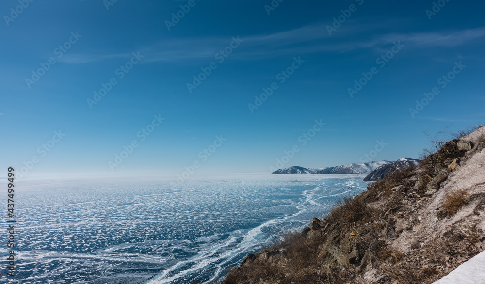 Cracks and lacy snow patterns are visible on the surface of the frozen lake. Blue ice and azure sky. There are stones and dry grass on the slope of the coastal hill. Baikal. Cracks and lacy snow patterns are visible on the surface of the frozen lake. Blue ice and azure sky. There are stones and dry grass on the slope of the coastal hill. Baikal.