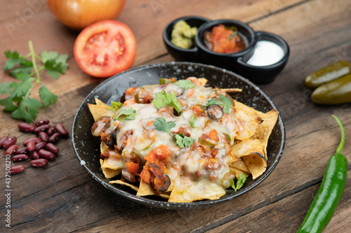 A plate of delicious corn chips nachos in cheddar cheese with beans, tomato, green chili, black olives, cilantro served with guacamole, sour cream and tomato salsa dip on the side isolated on the wood