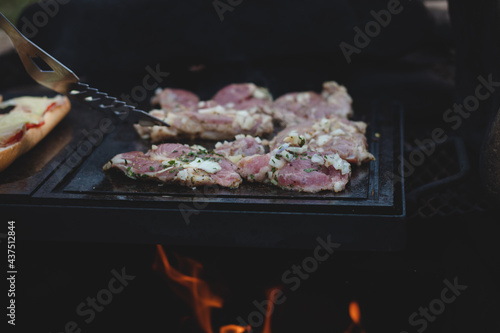 Man uses a steel ladle to turn a loaded pork neck with basil, salt, pepper and oil from one seared side to the other. Barbecue on a fierce outdoor fire. Barbecue season has begun