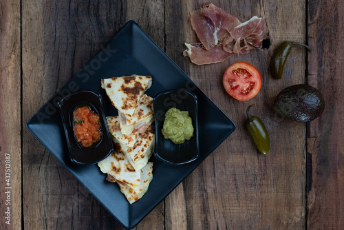 Top view of a toasted tortilla with chicken, peppers, red onion and mozzarella served with a guacamole dip and sour cream isolated on wooden table photo