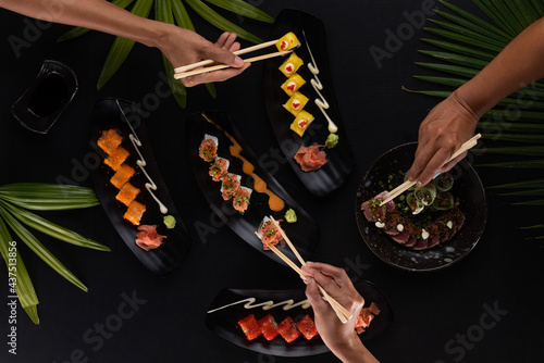 Top view hands with chopsticks and mix of sushi, Japanese food over black table with tropical leaves