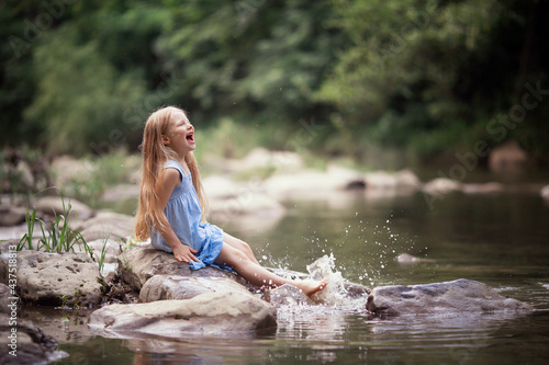 a little girl in the forest on the river sits on a rock splashing water