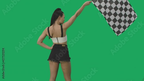 Rear view of brunette waving black and white checkered racing flags to signal start of competition. Young woman posing against background of green screen close up. Slow motion ready, 4K at 59.94fps photo