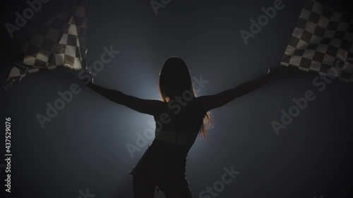 Rear view of silhouette young woman waving checkered race flag to signal the start of racing event. Brunette posing in a dark smoky studio with backlight. Close up. Slow motion ready, 4K at 59.94fps. photo