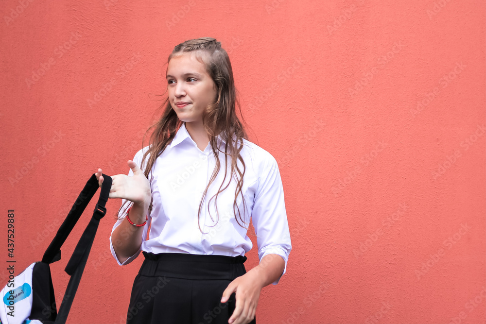 A cute beautiful teenage girl in formal school wear carrying a modern fashion backpack and standing against an orange wall. Back to school, education concept. Happy childhood background. Copy space.