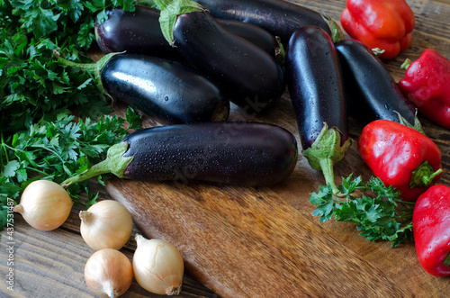 Whole raw eggplant, onions, red bell pepper and parsley on wooden cutting board.