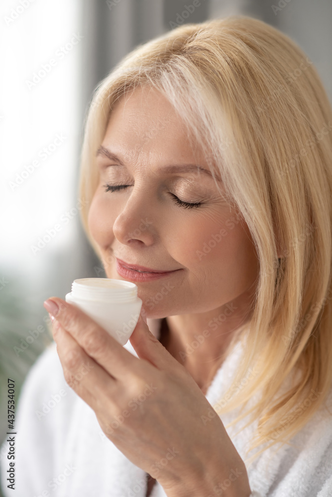 A woman in a white bath robe holding a jar of a facial cream and smelling it A woman in a white bath robe holding a jar of a facial cream and smelling it