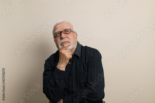 elderly gray-haired man in glasses and black shirt rubs pensively with his hand beard, emotions in the frame, waist portrait, light background, studio © mira_y