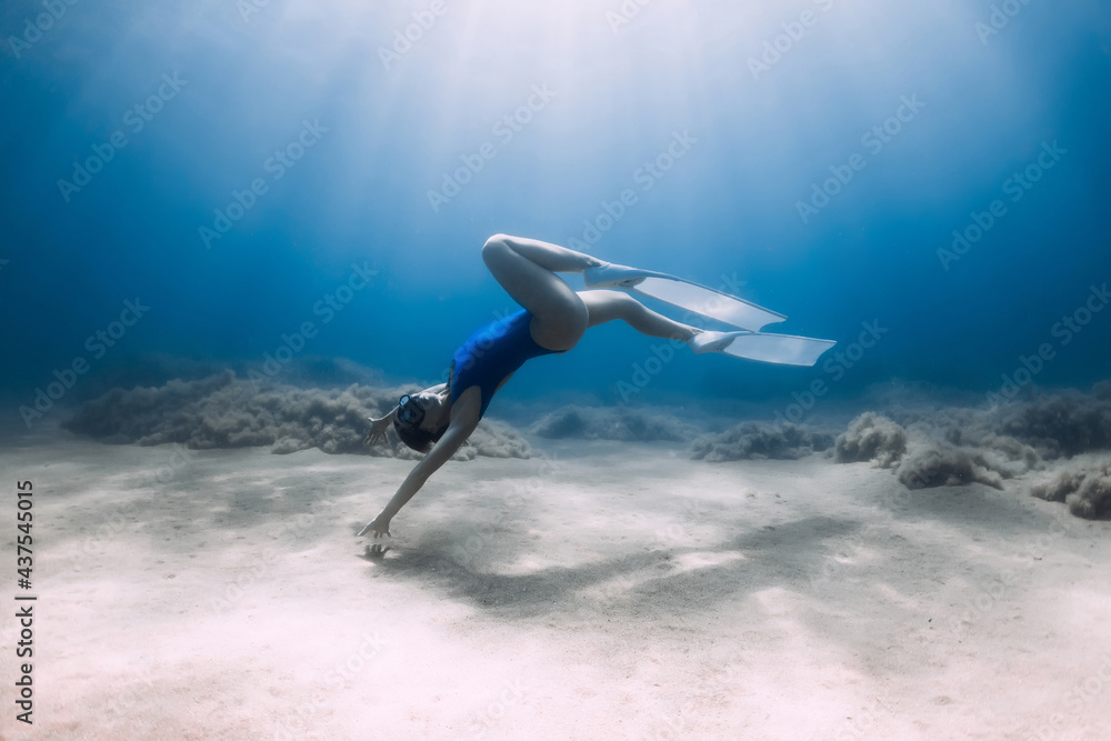 Lady freediver with fins posing and glides underwater in blue sea with sunlight.