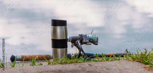 Fishing rod, spinning reel and a cup of hot coffee on the bank of the river pier. Out-of-town vacation concept. Selective focus