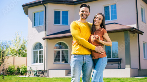 Modern beautiful excited young couple in love standing in front of the new big house with the keys in hands and hugging while they celebrating this buying