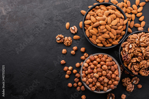 Assorted nuts. Top view of different types of peeled nuts in bowls and a few nuts scattered around on a black textured background, copy space.