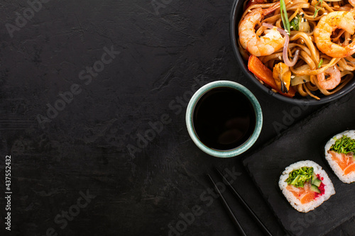 Wok noodles with shrimps and vegetables and sushi rolls on a black slate background, top view, copy space. Traditional Asian food set.
