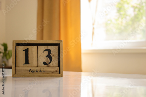 Wooden blocks of the calendar represents the date 13 and the month of April on the background of a window, curtain and a plant.