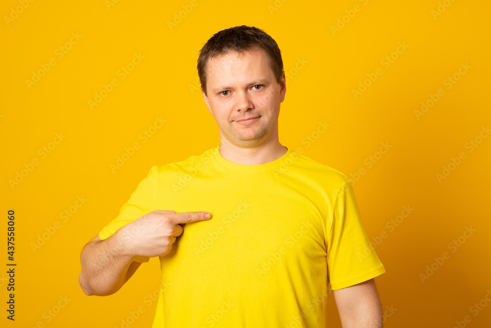 T shirt design and advertising concept. Style and fashion. Indoor shot of smiling man pointing index finger at copy space on her blank yellow top. T shirt design and advertising concept. Style and fashion. Indoor shot of smiling man pointing index finger at copy space on her blank yellow top.