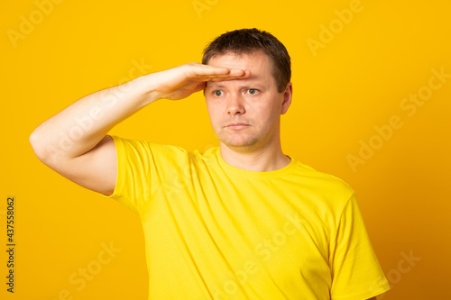 Young blond man wearing casual yellow shirt very happy and smiling looking far away with hand over head. Searching concept.