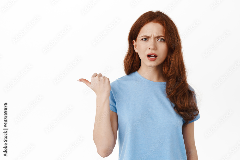 Confused and annoyed redhead girl pointing finger left, showing something strange and frustrating, cant understand what is that aside, standing in t-shirt against white background Confused and annoyed redhead girl pointing finger left, showing something strange and frustrating, cant understand what is that aside, standing in t-shirt against white background