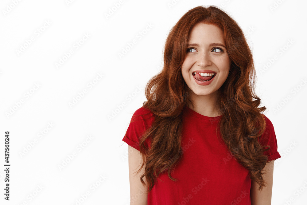 Portrait of happy pretty woman with red curly hairstyle, smiling thoughtful, lick lips from temptation to taste something delicious, looking left at copyspace, standing against white background Portrait of happy pretty woman with red curly hairstyle, smiling thoughtful, lick lips from temptation to taste something delicious, looking left at copyspace, standing against white background