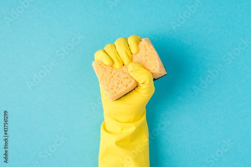 Top view photo of raised hand in yellow rubber glove clenching yellow scouring pad on isolated pastel blue background with copyspace photo