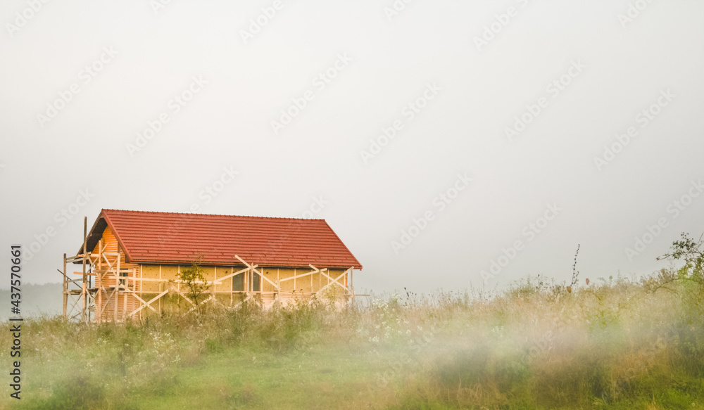 Morning landscape with construction of new wooden cottege house for tourists in the Carpathian mountains, chalet style building with the foreground of meadow covered by fog.